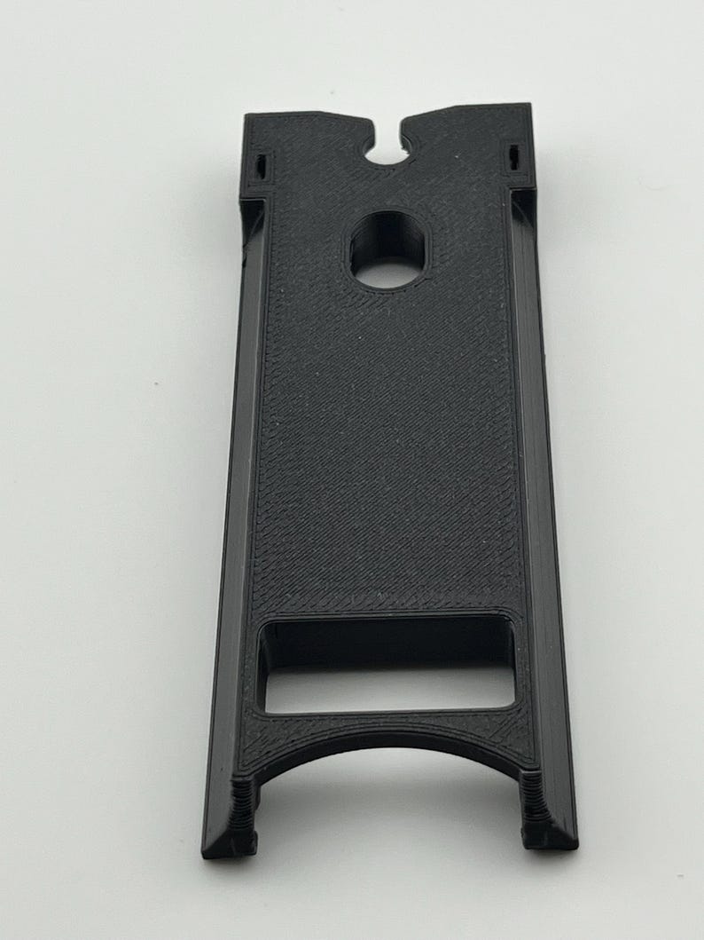 Puede incluir: Un objeto negro rectangular impreso en 3D con una superficie texturizada. Presenta un agujero circular cerca de la parte superior y una abertura curva en la parte inferior. El dise&ntilde;o incluye una ranura en la parte superior y una abertura rectangular.