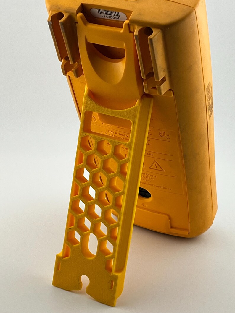 May include: A yellow digital multimeter with a built-in stand. The stand has a honeycomb design. The multimeter has a label with the number "11440054".