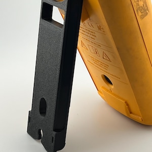 Puede incluir: Un soporte negro impreso en 3D que sostiene un mult&iacute;metro amarillo. El soporte es rectangular con una superficie texturizada y un recorte para la pantalla del medidor. El mult&iacute;metro tiene una etiqueta con texto y s&iacute;mbolos.