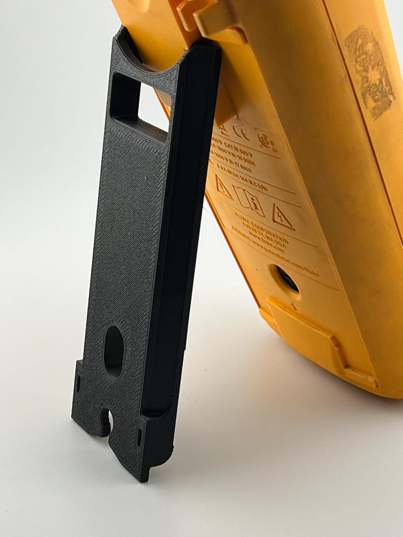 Puede incluir: Un soporte negro impreso en 3D que sostiene un mult&iacute;metro amarillo. El soporte es rectangular con una superficie texturizada y un recorte para la pantalla del medidor. El mult&iacute;metro tiene una etiqueta con texto y s&iacute;mbolos.