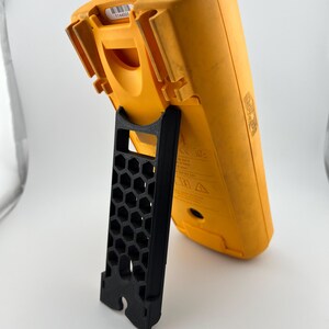 May include: A yellow digital multimeter with a black stand. The multimeter is angled, showing its back and side. The stand is black and has a honeycomb pattern.