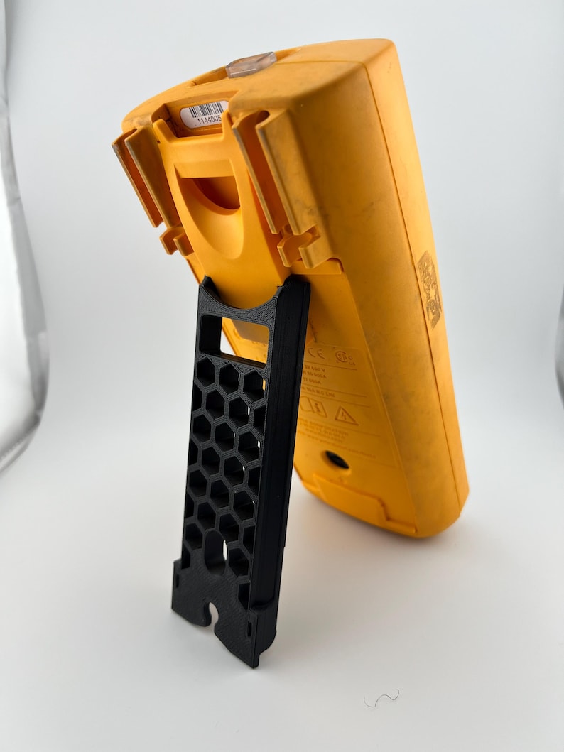 May include: A yellow digital multimeter with a black stand. The multimeter is angled, showing its back and side. The stand is black and has a honeycomb pattern.