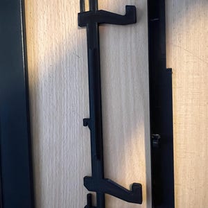 May include: A black plastic wall mount with two hooks and a long, narrow, vertical piece. The mount is designed to hold a picture frame or other object.