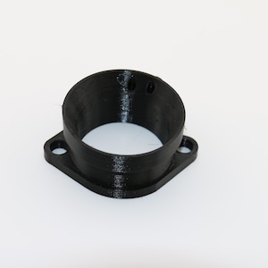 Op de afbeelding: Een zwarte, 3D-geprinte cilindrische flensadapter met een ronde opening en een vierkante basis met twee ronde montagegaten aan weerszijden. Het onderdeel heeft een gladde, matte afwerking en een zichtbare gelaagde textuur van het printproces.