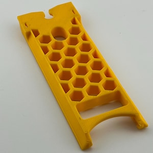 May include: A bright yellow, rectangular object with a honeycomb pattern and a handle. The object has a circular hole and a notched top. The design suggests a tool or a component for a larger device.