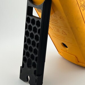 May include: A black 3D-printed stand supporting a yellow electrical testing device. The stand features a honeycomb pattern and a curved top to cradle the device. The device has printed text and is angled for viewing.