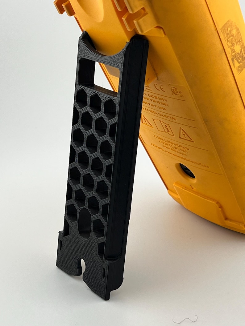 May include: A black 3D-printed stand supporting a yellow electrical testing device. The stand features a honeycomb pattern and a curved top to cradle the device. The device has printed text and is angled for viewing.