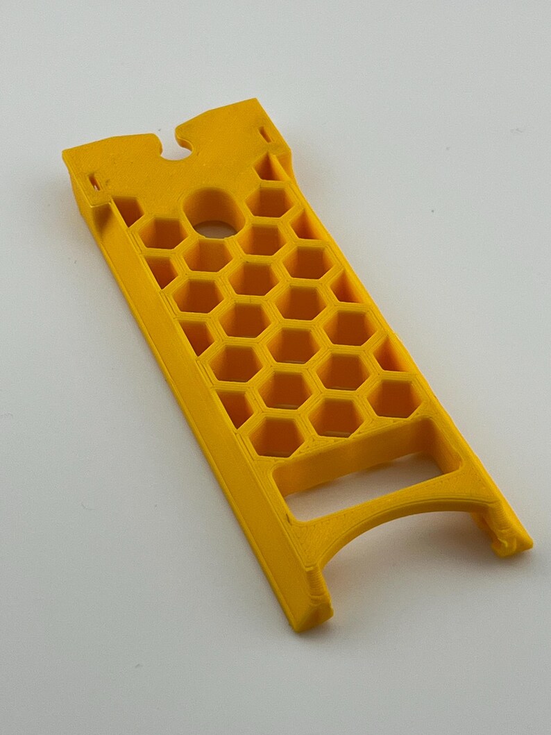 May include: A bright yellow, rectangular object with a honeycomb pattern and a handle. The object has a circular hole and a notched top. The design suggests a tool or a component for a larger device.