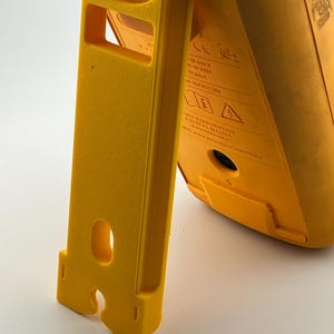 Puede incluir: Un mult&iacute;metro digital amarillo con un soporte integrado. El soporte tambi&eacute;n es amarillo y tiene forma rectangular con un recorte. El mult&iacute;metro tiene texto y s&iacute;mbolos impresos en su superficie. El dispositivo est&aacute; dise&ntilde;ado para mediciones el&eacute;ctricas.