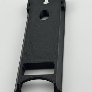 Puede incluir: Un objeto negro rectangular impreso en 3D con una base curva, un agujero circular y una muesca en la parte superior. El objeto tiene una superficie texturizada y parece ser un soporte o un soporte.