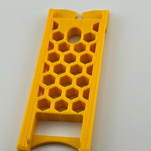 May include: A bright yellow, 3D-printed object with a honeycomb pattern. The object has a rectangular shape with a curved base and a hole in the center. The design features a series of hexagonal cutouts, creating a lattice-like structure.