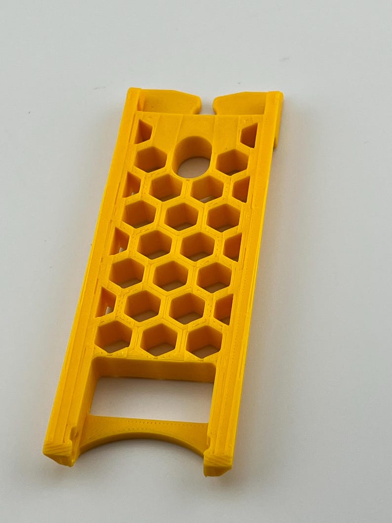 May include: A bright yellow, 3D-printed object with a honeycomb pattern. The object has a rectangular shape with a curved base and a hole in the center. The design features a series of hexagonal cutouts, creating a lattice-like structure.
