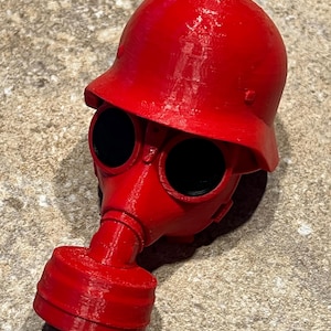 May include: A red gas mask with a red helmet. The gas mask has two large round eye pieces and a round filter.