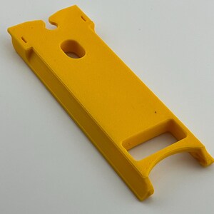 Puede incluir: Una herramienta de pl&aacute;stico rectangular de color amarillo brillante, con un mango curvo y un agujero central. La herramienta tiene una ranura en la parte superior y est&aacute; dise&ntilde;ada para un prop&oacute;sito espec&iacute;fico.