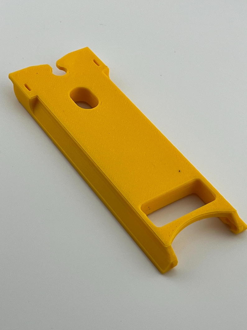 Puede incluir: Una herramienta de pl&aacute;stico rectangular de color amarillo brillante, con un mango curvo y un agujero central. La herramienta tiene una ranura en la parte superior y est&aacute; dise&ntilde;ada para un prop&oacute;sito espec&iacute;fico.