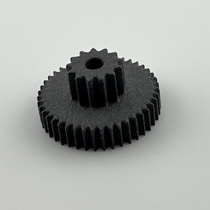 Peut inclure: Une roue dentée noire imprimée en 3D, avec un trou central et deux rangées de dents. La roue est posée sur un fond blanc uni, soulignant son design mécanique et sa texture. La roue mesure environ 5 cm de diamètre.
