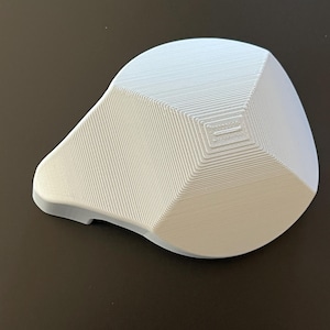 May include: A white, 3D-printed object with a geometric design. The object has a rounded top and a flat base, with a series of parallel lines creating a textured surface. The object is likely a decorative item or a functional piece.