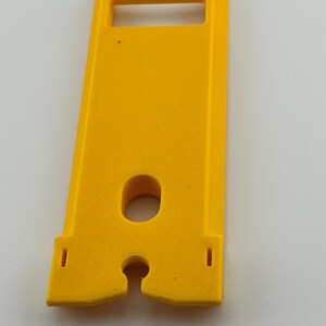 Puede incluir: Un componente de pl&aacute;stico amarillo brillante con forma rectangular, con una abertura tipo asa en la parte superior y un agujero circular en el centro. Dos peque&ntilde;os recortes rectangulares en la parte inferior. Probablemente parte de un mecanismo m&aacute;s grande.