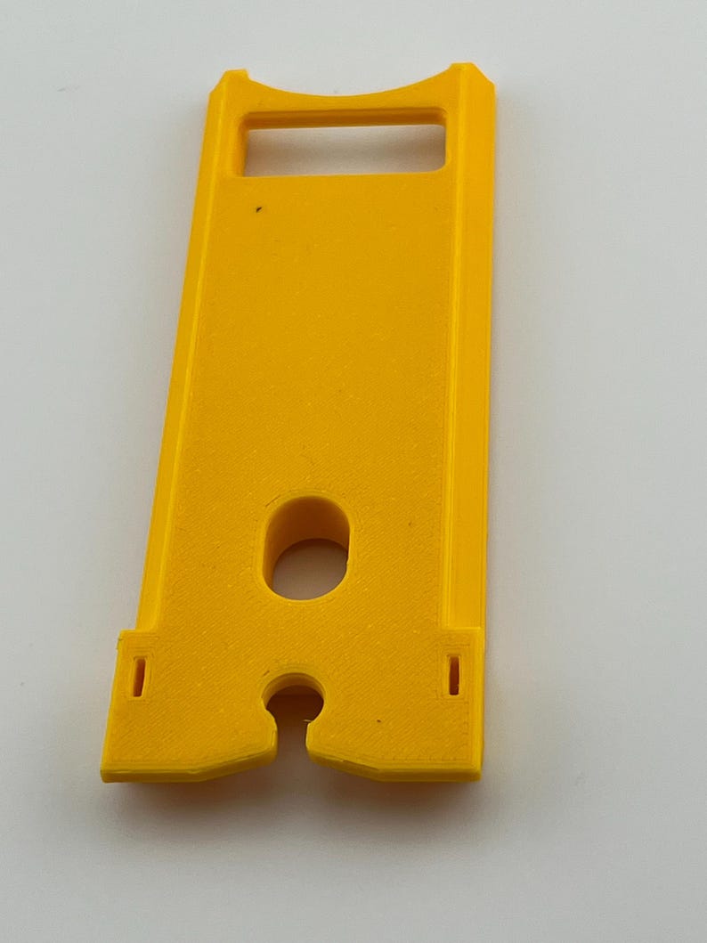 Puede incluir: Un componente de pl&aacute;stico amarillo brillante con forma rectangular, con una abertura tipo asa en la parte superior y un agujero circular en el centro. Dos peque&ntilde;os recortes rectangulares en la parte inferior. Probablemente parte de un mecanismo m&aacute;s grande.