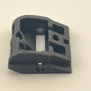 Puede incluir: Una pieza de plástico negro impresa en 3D con una abertura rectangular y varios agujeros. La pieza está inclinada y tiene una superficie lisa.