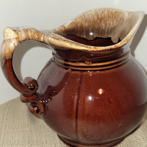 May include: A brown ceramic pitcher with a rounded body and a wide, flared rim. The handle is a lighter brown color with decorative swirls. The pitcher has a glossy finish and appears to be vintage.