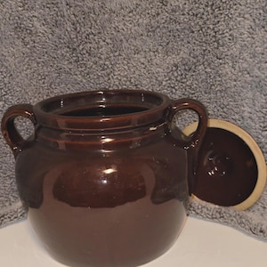 May include: A brown ceramic crock with two handles and a lid. The crock is glazed and has a smooth finish.