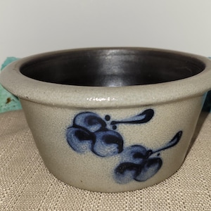May include: A stoneware bowl with a light beige exterior and a dark interior. The bowl features a blue floral design. The rim is rounded, and the bowl sits on a textured surface. A decorative piece for the kitchen.