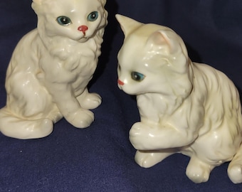 May include: Two white ceramic cat figurines. One cat is sitting upright, the other is in a playful pose. Both have blue eyes and red noses. The figurines are on a dark blue background.