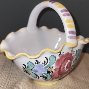May include: A white ceramic basket with a handle, featuring a scalloped rim and floral designs. The handle is decorated with yellow and purple stripes. The basket has a yellow base and is decorated with red, blue, and green flowers.