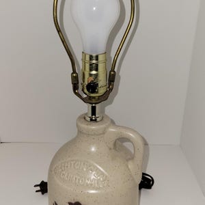 May include: A vintage stoneware lamp with a cream-colored jug base featuring a handle and a floral design. The lamp has a brass-toned frame, a white bulb, and a dark brown finial. The base has the text "CLINTON" on it.
