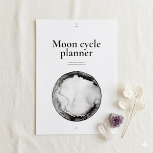 May include: A white Moon Cycle Planner with black text and a watercolor-style moon illustration. The planner includes the text "Work with your intentions in rhythm with the lunar cycle." A clear quartz crystal, amethyst crystal, and dried plant are next to the planner.