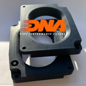 Może przedstawiać: Dwa czarne plastikowe rozpórki kolektora dolotowego z nadrukiem "DNA HIGH PERFORMANCE FILTERS" w kolorze pomarańczowym.