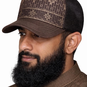 Puede incluir: Gorra de camionero marrón con un panel frontal estampado y una parte trasera de malla negra. El panel frontal presenta un diseño geométrico repetitivo en un color marrón más claro. La gorra tiene una visera curva.