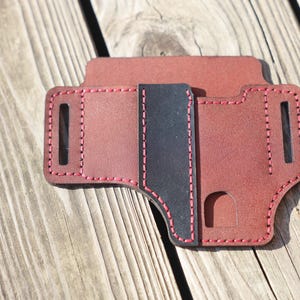 May include: A handmade leather holster in shades of brown and black, with red stitching. The holster is designed to hold a small item and features a belt loop. The item is placed on a wooden surface.