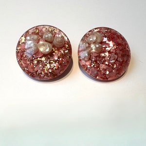 Op de afbeelding: Een paar roze en gouden glitteroorbellen met witte schelpen ingebed in de hars.
