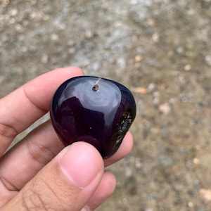 May include: A polished, dark purple gemstone with a smooth, reflective surface. The stone has a small, drilled hole near the top and a slightly rough area on one side. The gemstone is held in a hand.