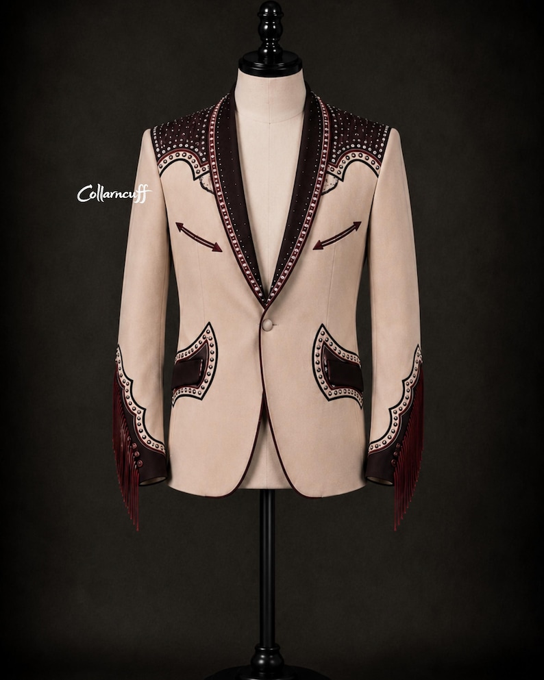 May include: A beige blazer with dark brown accents and decorative stitching. The blazer features fringe on the sleeves and is displayed on a mannequin. The collar and pockets are trimmed with dark brown fabric.