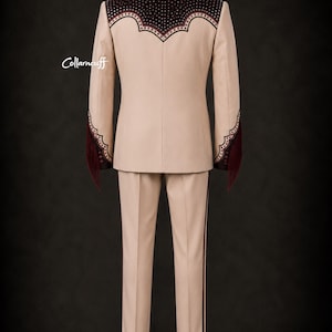 May include: A beige suit with decorative burgundy accents. The jacket features a detailed yoke and sleeve cuffs with fringe. The pants have a burgundy stripe down the side. The suit is displayed against a dark background.