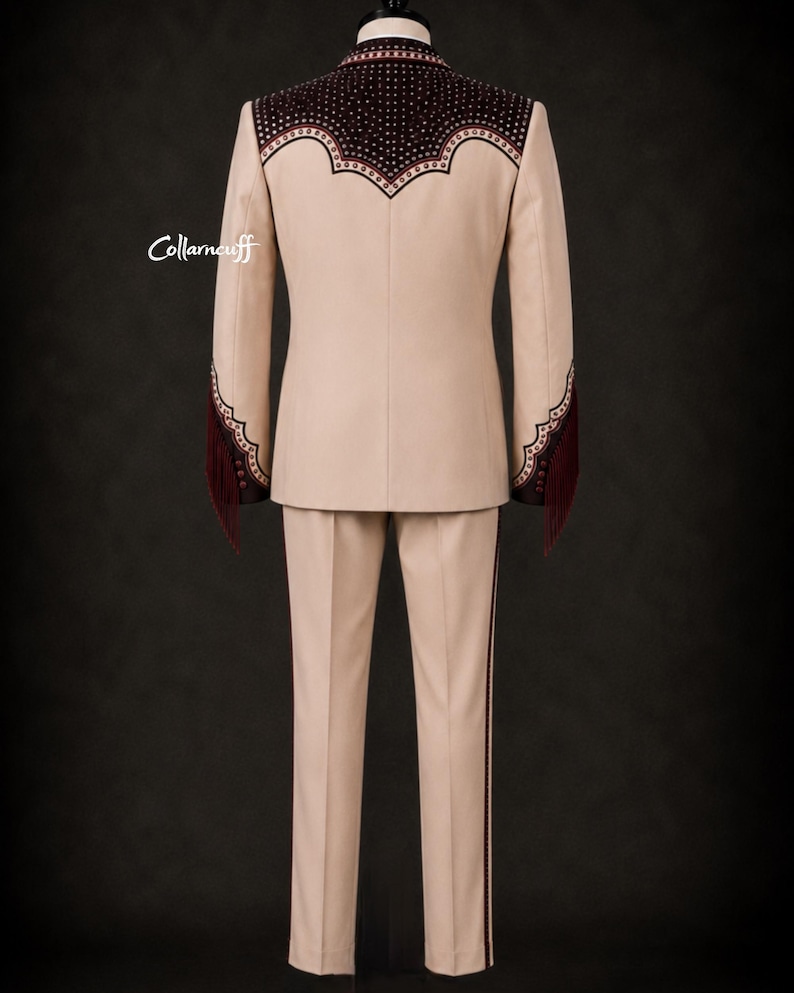 May include: A beige suit with decorative burgundy accents. The jacket features a detailed yoke and sleeve cuffs with fringe. The pants have a burgundy stripe down the side. The suit is displayed against a dark background.