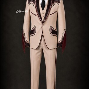 May include: A tan suit with decorative dark brown accents, including fringe on the sleeves. The suit features a blazer, vest, and trousers, with a black tie and white shirt. The suit has decorative stitching and trim.
