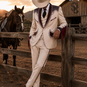 May include: A cream-colored Western-style suit with burgundy accents, worn with a matching hat and black tie. The suit features decorative stitching and fringe details. The scene is set outdoors with a horse and a wooden fence in the background.