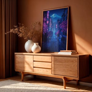 May include: A wooden dresser with three drawers and two doors. The dresser is light brown and has a modern design. A framed print of a city street scene with a mountain in the background is on top of the dresser. The print is blue and purple with pink accents.