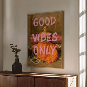 Könnte beinhalten: Ein gerahmter Druck eines Gemäldes, das eine Frau in einem langen orangenen Kleid zeigt, die auf einem Sofa sitzt. Die Frau liest ein Buch und hat eine rosa Blase im Mund. Der Text "GOOD VIBES ONLY" ist in rosa Buchstaben über dem Gemälde geschrieben.