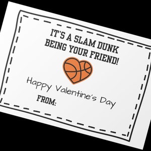 Peut inclure: Une carte blanche avec une bordure noire et un cœur en forme de ballon de basket. La carte indique "It's a slam dunk being your friend!" et "Happy Valentine's Day". Il y a un espace pour écrire "From:"