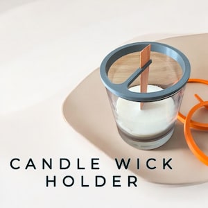 Può includere: Un porta stoppino per candele grigio con uno stoppino di legno all'interno. Il porta stoppino è in plastica e ha una base in vetro trasparente. Il porta stoppino è su una superficie beige. "CANDLE WICK HOLDER" è scritto in nero sotto il porta stoppino.