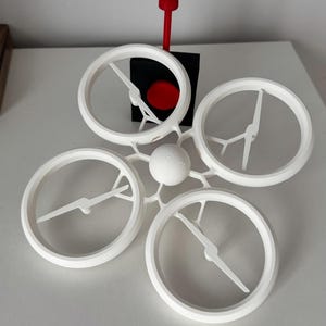 Può includere: Un drone bianco con quattro eliche e una leva di controllo rossa. Il drone è su una superficie bianca.