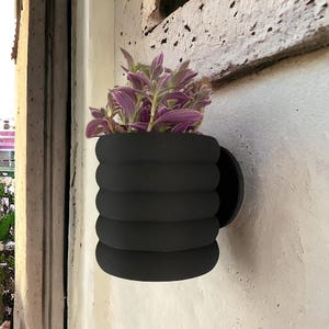 May include: A black, ribbed, wall-mounted planter with a purple and green plant inside.
