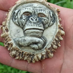 May include: A carved monkey face pendant made from antler. The monkey is depicted with a serious expression and its hands are clasped together. The antler is a natural brown color with a rough texture.