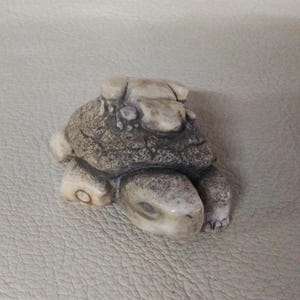 Op de afbeelding: Een kleine, gedetailleerde schildpadfiguur in grijstinten en beige. De schildpad heeft een getextureerd schild en zichtbare poten en kop. Het beeldje is waarschijnlijk gemaakt van steen of een vergelijkbaar materiaal.