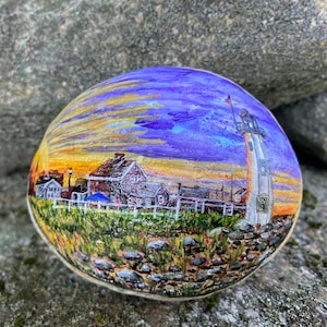 May include: Hand-painted rock featuring a coastal scene with a lighthouse, buildings, and a sunset sky. The artwork depicts a white lighthouse with a red-roofed building and a white picket fence. The rock is resting on a gray stone surface.